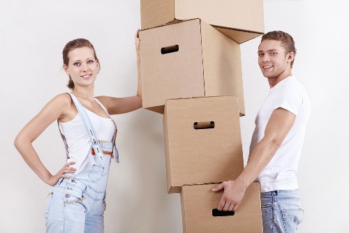 A young man and woman, both smiling, are indoors in a residential setting with plain white walls. The woman, dressed in light denim overalls and a white tank top, stands on the left with her right hand on her hip. The man, wearing a white t-shirt and light jeans, is on the right, holding a large cardboard box in his left hand, with additional boxes stacked in between them. The boxes are plain brown with black handles, varying in size, and are arranged vertically, suggesting they are in the process of being moved or packed. The ambient lighting is bright and even, typical of a well-lit hallway or living room area. The scene subtly reflects the context of house removals or furniture relocation, with no visible furniture or household items apart from the boxes, emphasizing the focus on packing. The natural setting and professional demeanor of the individuals align with a house moving service typical of a company like Watson Removals London servicing areas such as Arkley, with attention to careful handling and organization during a residential move.