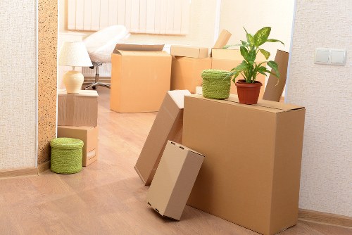 A residential interior with a collection of unpacked cardboard moving boxes of varying sizes placed on and around a wooden floor. Several boxes are upright, some are lying on their sides, showing plain brown surfaces. On top of one large box, there's a small potted plant with broad green leaves and a textured woven green container. Nearby, a smaller round green container is positioned on the floor, leaning against a box. In the background, a white table lamp with a shade and a small decorative object can be seen, along with a bed partially visible in a bedroom area. The room has light-colored walls and a subtle textured wallpaper, with a neutral, soft atmosphere created by natural lighting. This scene appears to capture a moment during a house move or relocation within a London suburb, reflecting typical household packing activity undertaken by Watson Removals London in the area code post-designation.