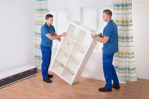 Two male furniture movers dressed in blue work uniforms are in a well-lit room with white walls and a wooden floor. They are carrying a white modular shelving unit with multiple compartments, positioning it carefully between them as part of a house removal process. The room features a large window with white blinds and multicoloured striped curtains, allowing natural light to illuminate the scene. In the background, there is a black and white radiator along the base of the wall. The environment appears to be a residential living space, possibly an apartment or house in West Brompton, with a clean and tidy appearance. The movers are focused on handling the heavy item securely, using proper lifting techniques to avoid damage. The setting suggests a professional removals service by Watson Removals London, experienced in handling household furniture and belongings during relocations within London postcodes such as SW10. The furniture's smooth surface and white finish contrast with the warm tone of the wooden floor and the casual, yet practical attire of the movers, emphasizing careful handling of household items typical of house removals in West Brompton.