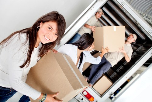 In this image, three professional movers are engaged in loading a residential removals van, likely in a suburban area near London, as suggested by the context of Watson Removals London. The foreground features a smiling woman with long brown hair, wearing a white top, carrying a medium-sized cardboard box with no visible markings, textured with a corrugated surface. Behind her, a woman with dark hair, dressed in a similar white top, is reaching into the van, holding a box while standing on the vehicleâ€™s lower lip. To the right, a man with short light-colored hair, dressed in a neutral-toned shirt and dark trousers, is standing inside the van with the door open, holding a larger cardboard box. The van's interior shows metallic ridges and a partially visible load area, with additional boxes stacked nearby. The scene is set outdoors during daylight, with natural light illuminating the group and their equipment, emphasizing the professional and organized nature of a house move by Watson Removals London as they prepare for a residential relocation within the London boroughs or surrounding postcode areas in the UK.