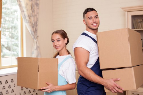 A young man and a young woman, both smiling, are inside a well-lit residential room with cream-colored walls and a window with patterned curtains in the background. The man, wearing a white t-shirt and navy blue apron, is holding two cardboard boxes stacked on top of each other in his hands, with the larger box on top. The woman, dressed in a light blue dress with white sleeves, is holding a single medium-sized cardboard box, supporting it with both hands. Behind them, a kitchen unit with open shelving is partially visible. The room appears to be preparing for a house move, with the individuals engaged in packing or unpacking. The environment emphasizes the domestic setting typical of house removals, with a focus on the cardboard boxes, the individuals' attire suited for manual handling, and the natural light streaming from the window from outside in London, subtly connecting to the local context of residential house relocation services provided by Watson Removals London, covering areas like the postcode inferred from the page title and URL. The overall scene conveys organized and professional household moving activities.