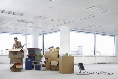 The image depicts a person in a business office environment engaged in packing or organizing belongings for a house move, within a spacious room filled with natural light from large windows revealing a cityscape background. They are wearing a patterned shirt and are leaning over a cardboard box, handling items or preparing packing materials. Surrounding them are various cardboard boxes of different sizes, some sealed and others open, with packing paper or other materials visible inside. Several boxes are stacked or placed on the floor, while others are arranged on a desk or table area. An office chair is placed nearby, and electrical cords are seen on the floor, possibly connected to electronic devices or packing equipment. The room features a plain white ceiling with ceiling-mounted fluorescent lights, and the walls are painted in a light neutral tone. The overall scene emphasizes the process of organizing personal belongings for a move, related to residential relocations near Cranham, with Watson Removals Londonâ€™s services implied through the context of house removal preparations.