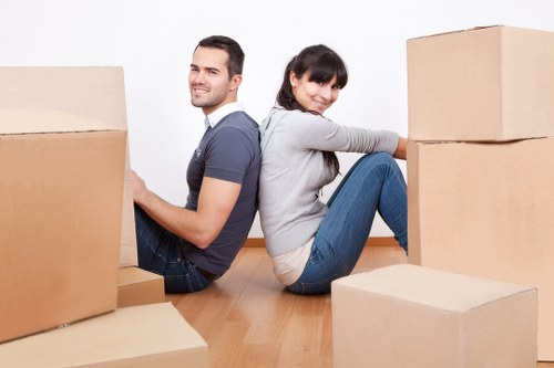 A young man and woman sit back-to-back on a wooden floor in a bright residential interior, surrounded by several large cardboard moving boxes in various stages of packing. The man, dressed casually in a navy t-shirt and jeans, is smiling and looking slightly to his right, while the woman, wearing a light grey sweater and jeans, is also smiling and looking towards the camera. The boxes are positioned around them, some stacked and others placed singly, with the larger boxes behind them and a smaller box in the foreground. The scene reflects the process of house relocation, with a clean, well-lit environment suggesting a home setting in London or the surrounding area. Watson Removals Londonâ€™s professional domestic moving service is implied by the scene, which captures a calm and organized moment during a house move or packing activity, emphasizing careful handling and preparation typical of local removal specialists.