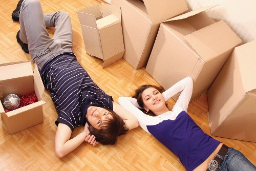 Two young adults, a male and a female, are lying on a wooden floor inside a residential property surrounded by open cardboard moving boxes of various sizes. The male, dressed in a navy and white striped T-shirt and grey trousers, is lying on his back with his head near a box that contains decorative ceramic balls, suggesting they are in the process of packing or unpacking belongings. The female, wearing a white top with a blue pinafore dress, is lying on her back with her hands behind her head, smiling and looking relaxed. The boxes around them are partially filled and sealed, indicating an ongoing house move or packing activity typical of a house removals service. Natural lighting illuminates the scene, emphasizing the casual and comfortable atmosphere of a residential interior, possibly in Ruxley, London, during a relocation process facilitated by Watson Removals London.