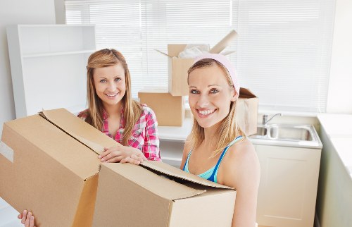 Two women, likely a moving team or homeowners, are indoors in a well-lit, modern kitchen with white cabinetry, a stainless steel sink, and a window with closed blinds. They are holding a large, partially opened cardboard box, which appears to contain additional packing materials or household items. One woman, dressed in a pink and white checkered shirt, is smiling and standing behind the box, while the other woman, in a blue tank top and a light pink headband, is also smiling and positioned to the right. The background shows a tidy kitchen environment, with some stacked cardboard boxes on a countertop, suggesting they are engaged in packing or unpacking during a house move in Mill Hill. The scene reflects a professional and organized approach to a residential removal, consistent with services offered by Watson Removals London, with a focus on a smooth transition for clients. The overall atmosphere is friendly, efficient, and clean, emphasizing attention to detail in a typical house relocation setting within a London suburb.