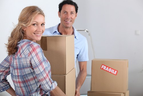 A smiling woman with blonde hair wearing a plaid shirt holds a cardboard box in the foreground, while a man with dark hair in a light blue shirt stands behind her, both appearing to be in the process of moving. In the background, there are additional cardboard boxes, one of which has a red 'Fragile' sticker, and a metal trolley is visible. The scene takes place inside a bright, modern house or apartment with a plain white wall and an open space typical of a residential setting in Yeading. Nearby, a ladder and minimal furniture suggest an active house removal or packing process conducted by Watson Removals London, a professional house removals company serving the area. The overall environment is clean and organized, with a focus on household items such as cardboard boxes, which are stacked and ready for transport, reflecting a typical domestic move.