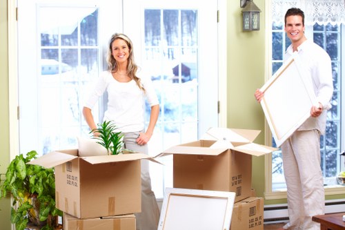 A smiling woman with long blonde hair, dressed in a white top and grey trousers, stands inside a bright residential room near a large window with white curtains, surrounded by open cardboard boxes used for packing. In front of her are several stacked boxes, some sealed and others open, revealing a potted green plant with broad leaves, alongside additional packing materials. To her right, a man in a white shirt and light-coloured trousers holds an empty picture frame, also standing near the window, which offers a view of a residential street typical of Lewisham with snow visible outside. The room features light green walls and a brass wall-mounted lantern beside the window. The scene appears to capture the process of preparing for a house move, with the household items and boxes arranged for packing and transportation, supported by Watson Removals Londonâ€™s local expertise in Lewisham removals services.