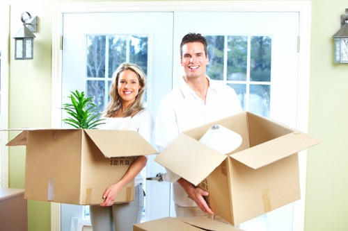 A smiling man and woman are standing inside a residential home in Marks Gate, holding open cardboard boxes filled with packing materials and a small potted plant. The woman on the left is dressed in a white top and gray trousers, and she holds a box containing a green potted plant, a roll of packing paper, and some other household items. The man on the right is wearing a white shirt and gray trousers, holding a larger box with additional packing supplies. Behind them, there are double-glazed patio doors with white frames, providing a view of a garden with green foliage. The interior walls are painted light green, and decorative wall-mounted lights flank the door. The scene depicts prepared household items ready for relocation, consistent with professional house removals services in London, specifically in the area of Marks Gate, showcasing the process of packing and organizing belongings for a move.