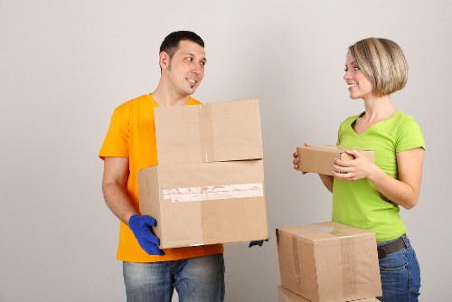 In a residential setting, a man wearing a bright yellow T-shirt and blue work gloves holds a cardboard box with a plain brown exterior and white packing tape across the middle, while a woman dressed in a lime-green T-shirt and jeans stands opposite, smiling as she receives a similar cardboard box. Behind them, a stack of additional boxes of varying sizes, all made of cardboard with minimal markings, rests on the floor, suggesting an ongoing moving process typical of house removals. The background features a plain, light-colored wall, and the scene is illuminated evenly, emphasizing the natural light within the interior space. The image conveys a professional, organized approach to relocating household items, aligning with Watson Removals London's services in Maze Hill, London, focusing on careful handling and packing of personal belongings during house moves in the local area.