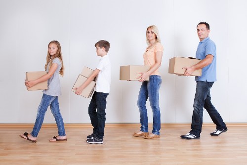 A family of four, consisting of two young children, a woman, and a man, are standing indoors in a well-lit room with a plain white wall and wooden floor. They are carrying cardboard boxes, which vary in size and are sealed with tape, indicating they are engaged in a household move. The children are at the front, each holding a medium-sized box, with the girl dressed in a grey top and blue jeans, and the boy in a white shirt and dark trousers. The woman, positioned behind them, is holding a slightly larger box, dressed in a peach top and blue jeans. The man is at the back, carrying a large box with both hands, dressed in a blue shirt and black trousers. The scene captures a moment of packing or unloading during a house removal, with a focus on the family safely handling their belongings. The environment appears to be a residential space, possibly a living room or hallway, with no additional furniture visible, emphasizing the household packing activity. The image subtly supports services like Hampton Hill removals by Watson Removals London, providing a clear depiction of a family participating in a home relocation process within the local area.