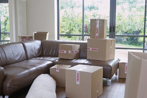 The image displays a living room with a dark brown leather sofa situated against large floor-to-ceiling windows that reveal a garden outside. Several cardboard moving boxes, labeled with red 'FRAGILE' stickers, are stacked on and around the sofa. The boxes vary in size and are packed with household items for moving, with some placed on the sofa cushion and others on the wooden floor. A light-colored fabric armchair is partially visible in the foreground. The natural daylight illuminates the room, highlighting the textures of the leather upholstery, the cardboard boxes, and the view of the greenery outside. The scene suggests a house in the process of being packed for a move, with the environment indicative of a residential property in Romford, consistent with the context of Watson Removals London's services for local house relocations.