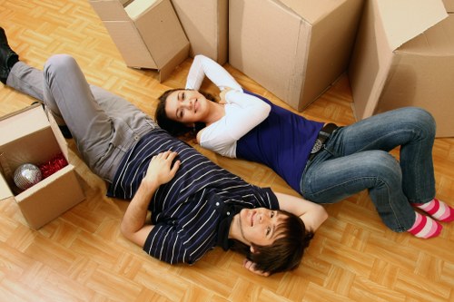 A man and a woman lie on their backs on a wooden-floored room surrounded by open cardboard moving boxes, some filled with household items such as decorative baubles and a rolled-up rug. The man is wearing a navy and grey striped T-shirt and light grey trousers, while the woman is dressed in a dark blue top and jeans with pink slippers, her head resting on her hands. The boxes vary in size and are positioned around them, with one containing a few plastic bottles and another with a rolled-up mat. The scene suggests a recent or ongoing house move, possibly in Stanmore, with the couple taking a break among packed belongings. The lighting appears natural, illuminating the room evenly, and the overall setting indicates an interior space preparing for or during a house relocation accessible to Watson Removals Londonâ€™s services in the local area.