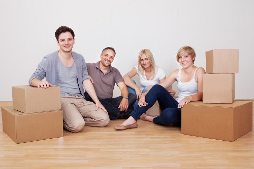 Four individuals, two men and two women, sit on a light wooden floor inside a room with a plain white wall, surrounded by various cardboard moving boxes of different sizes. The group appears relaxed and smiling, with some boxes stacked on their sides and others positioned upright. One of the men is wearing a casual grey long-sleeved shirt and beige trousers, while the other man is dressed in a grey polo shirt and dark pants. One woman has long blonde hair and wears a white top with dark jeans, and the other woman has short blonde hair and is dressed in a white tank top and blue jeans. The boxes are unsealed and appear ready for packing or unpacking, suggesting a house removal or relocation scenario typical of services provided by Watson Removals London in Dagenham. The environment indicates an indoor residential setting, emphasizing a professional yet casual atmosphere suitable for domestic moving services in the London area.