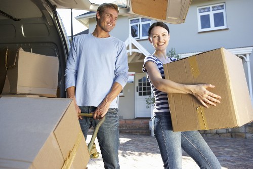 A man and a woman, both smiling, are engaged in loading a removals van outside a residential property in Arkley. The man, dressed in a light blue long-sleeve shirt and jeans, is holding a trolley with a large cardboard box on it, positioned on the driveway. The woman, wearing a striped top and jeans, is carrying a similarly large cardboard box with yellow packing tape, preparing to place it inside the van. The open back of the van reveals additional cardboard boxes and packing materials. In the background, the residential house features white-framed windows, a white porch, and a garden with a path and some shrubbery. The scene takes place during daylight with clear weather, indicating a typical household move in the Arkley area by Watson Removals London as part of their professional removals service. The environment suggests a peaceful suburban setting, with house exterior details and the logistics of packing and loading visible in the image.