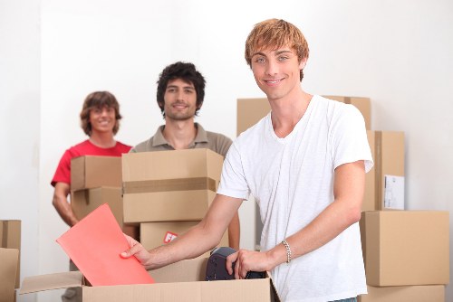 The image shows three young adults with light skin in a residential setting, engaged in a house move. The foreground features a young man with blond hair wearing a white T-shirt, smiling as he carries a medium-sized cardboard box with a pink envelope or folder placed on top, which he holds with one hand. Behind him, another young man with dark hair and a beard, dressed in a light-colored shirt, is holding a large cardboard box close to his chest, with a slight smile on his face. To the left, a young woman with shoulder-length brown hair, wearing a red T-shirt, stands smiling and holding a cardboard box close to her body. The background includes more cardboard boxes of varying sizes, some stacked, suggesting a moving process taking place within a home in Hampton Hill, as part of a professional removals service by Watson Removals London. The environment is well-lit, with a neutral interior setting, and the group appears to be in a spacious room or hallway ready for the next stage of their move, emphasizing a professional and organized household removals scene typical of a domestic move within London suburbs.