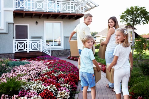 A family of four, consisting of a father and three children, are standing outside a residential house in Hampton Hill during daylight hours. The father, dressed in a light-colored shirt and jeans, is smiling as he holds a cardboard box, while the three childrenâ€”two girls and a boyâ€”are also holding boxes and looking towards the camera. The children are dressed casually, with one girl in a white top and trousers, and the boy in a light blue t-shirt and shorts, the other girl wearing a white dress. They are positioned on a paved pathway in front of a house with a modern exterior featuring white and grey elements, including a balcony with white railings and large windows. In the foreground, there is a well-maintained garden with vibrant pink, red, and white flowering plants, and green shrubbery lining the pathway. The background shows a residential neighborhood with neighboring houses, trees, and a bright, clear sky, suggesting a pleasant spring or summer day. Watson Removals London appears to be preparing for a house move, with the family engaging in packing or loading belongings amid the charming suburban setting of Hampton Hill, with the postcode area around TW12. The scene captures the active and organized process of a household relocation in a typical London suburb.