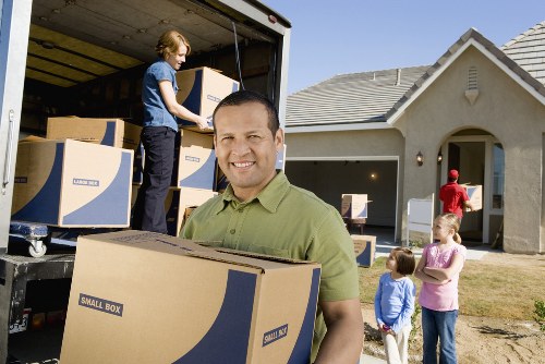 A professional house removal team is engaged in unloading cardboard boxes from a large removals van parked outside a modern detached house in a residential area, which has a light grey tiled roof and a cream exterior. One mover, wearing a green polo shirt, stands in the foreground holding a cardboard box labeled 'Small Box,' smiling at the camera. In the background, a female colleague in a blue shirt is positioned inside the van, handling more boxes, while three children, two girls in pink and purple tops and a boy in a blue shirt, stand nearby on the driveway observing the activity. The scene takes place during the daytime with clear weather, and the driveway is clean and paved, bordered by a well-maintained lawn and garden. The image captures a typical unpacking process during a house move in White City, illustrating efficient and organized unloading of household items by Watson Removals London, emphasizing reliable local moving services.