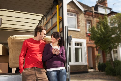 A young man and woman stand close together at the open back of a removals van parked outside a residential property in Leaves Green. The man is wearing a red long-sleeve shirt and beige trousers, while the woman is dressed in a purple cardigan over a white top and blue jeans. They are smiling at each other and appear to be engaged in conversation, with the woman holding a clear glass mug in her hand. Inside the van, there are cardboard boxes and a large beige sofa, indicating that they are in the process of moving house. The background shows a typical British terraced house with brickwork and white window frames, along with some greenery including a tree and bushes. Natural daylight illuminates the scene, suggesting a clear and calm day. This scene is representative of the residential moving services provided by Watson Removals London in the Leaves Green area, with household furniture and personal belongings ready for transport and relocation.