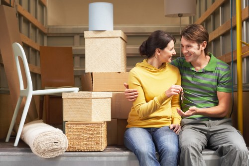 A young couple sitting on the floor of a moving storage unit or van, surrounded by household moving supplies and packed boxes. The woman, wearing a mustard yellow top, is smiling and holding a glass, while the man, dressed in a green and navy striped shirt, is smiling back and looking at her. Behind them, there are stacked cardboard boxes of various sizes, some with smooth surfaces and others with textured wrapping, alongside a wicker storage basket, a rolled-up cream-colored carpet, and a white plastic chair with a curved backrest. The background features wooden shelving and a cardboard box with a segment of a blue lampshade visible on top, indicating a household environment or a relocation setting. The lighting is even, highlighting the scene's warm and friendly atmosphere typical of a house move in the London area, supported subtly by the presence of typical removal supplies and the setting within a storage or moving vehicle.