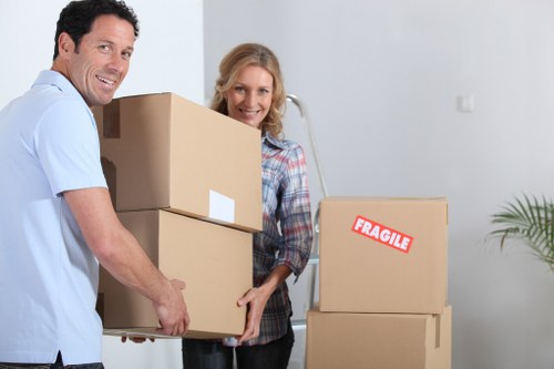 A man and woman, both smiling, are inside a residential setting with white walls, working together to carry three cardboard moving boxes. The man, dressed in a light blue shirt, holds a stack of two boxes, one on top of the other, with white labels visible on the boxes. The woman, wearing a plaid shirt, assists by supporting the lower box and is positioned behind the man. To their right, there are additional cardboard boxes, one of which has a red 
