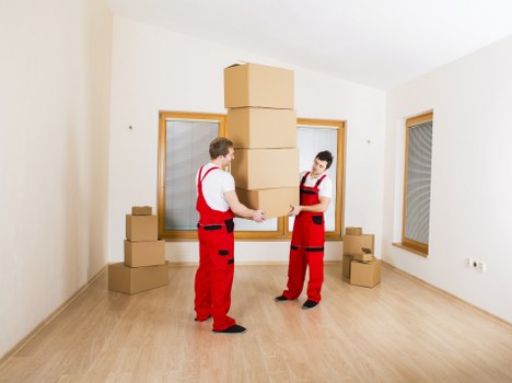 Two removal workers from Watson Removals London, dressed in red uniforms with black accents, are inside a bright, empty room with light wooden flooring and white walls, engaged in stacking and balancing cardboard boxes. One worker is holding a large box, while the other appears to be assisting in stabilizing the stacking process. Several additional cardboard boxes are arranged along the back wall, some stacked on the floor and others positioned near the windows, which have wooden frames. The room features two large windows, one on each side, allowing natural light to illuminate the scene, with suburban residential surroundings faintly visible through the windows. The environment looks clean and organized, reflecting a typical house removal or moving process taking place in Sidcup, with the focus on carefully transferring household items in preparation for relocation. The presence of a Watson Removals London team executing a professional and careful handling of boxes illustrates their expertise in house removals services in the area.