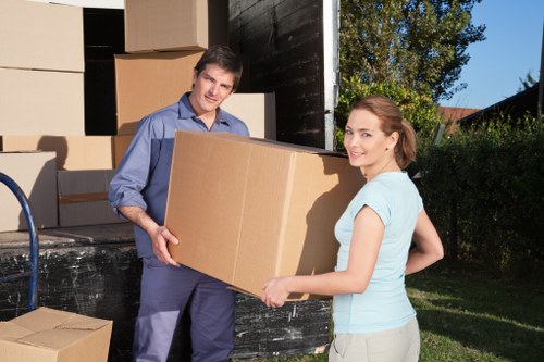 A young male and female mover, both dressed in casual clothing, are standing outdoors in a residential area near a house with a garden. The male mover, wearing a blue shirt and dark trousers, is holding a large cardboard box and smiling at the camera, while the female, wearing a light blue top and beige trousers, is assisting by supporting the box from the side. Behind them, an open moving truck with a dark exterior is visible, filled with stacked cardboard boxes of various sizes, some partially opened or closed. The background features greenery with trees and bushes, indicating a suburban setting, and the scene is illuminated by natural daylight, suggesting a daytime move. The environment appears clean and organized, with the movers engaged in loading or unloading belongings as part of a house removal process, typical of local relocations within the London area or nearby regions, showcasing the professional handling of household items.