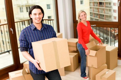 Two individuals are inside a bright, well-lit room with large windows that reveal a residential street in Clapham in the background. The person on the left, a young man with dark hair, is smiling and holding a medium-sized cardboard box wrapped with packing tape, dressed in a striped blue shirt. The person on the right, a woman with blonde hair, is also smiling and is positioned near a stack of cardboard boxes of various sizes, some open and some closed, ready for moving. She is wearing a red long-sleeve top and jeans, and appears to be in the process of packing or unpacking a box. The floor is light-colored and smooth, and the room's interior suggests a residential space suitable for house removals. Several boxes are arranged on the floor and against the windows, indicating an active packing or moving process typical of a professional removals service in Clapham. The scene captures a moment of preparation, with the Watson Removals London team engaged in an efficient and organized house moving task.