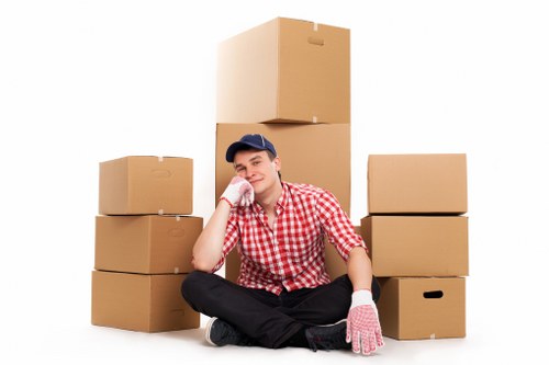 A young man sitting cross-legged on a white background, surrounded by several stacked cardboard boxes of varying sizes. He is wearing a red and white checkered shirt, black trousers, white gloves, and a dark blue cap. The boxes are plain, light brown with some featuring handles or open slots. The scene appears to represent a house removal or packing process, with no other furniture or environmental details visible. The man has a relaxed expression, supporting the idea of a professional yet approachable removals service. This image is suitable for a house removals website serving areas like Enfield, highlighting packing and moving activities undertaken by companies such as Watson Removals London. The focus remains primarily on the boxes and the individual, emphasizing the logistic aspect of a smooth move within or near London neighborhoods.