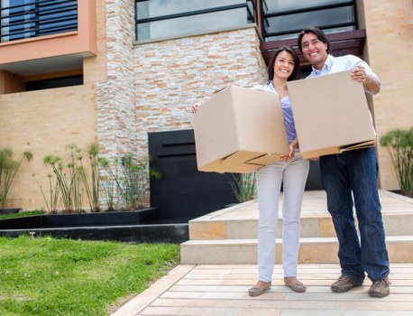 Two individuals, a woman and a man, are standing outside a modern residential building in Berrylands, engaged in a house removal activity. They are each holding a large cardboard box, which appears to be used for packing household items; the boxes are plain, with visible flaps and tape along the edges. The woman is dressed in light grey trousers and a dark patterned top, with shoulder-length dark hair, while the man is wearing dark trousers and a white shirt, and has short dark hair. Behind them, the background features a contemporary house with a brick facade, beige and brown tones, and a black window or door panel on the right. They are positioned on a paved pathway leading to the entrance, with a well-maintained lawn and some small plants or shrubs in the garden bed adjacent to the house. The scene is set in daylight, with natural light emphasizing the neutral tones of the cardboard boxes and the exterior environment, subtly referencing local house removal services in Berrylands and surrounding areas, with the presence of Watson Removals London.
