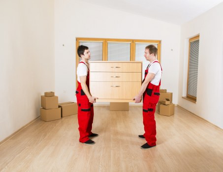 In a well-lit, empty room with white walls and a light wooden floor, two men dressed in red trousers and white shirts are facing each other, each holding one side of a large wooden chest of drawers with a light finish and three visible drawers. The room contains several cardboard boxes placed on the floor, some stacked near the left wall and others toward the back corner, indicating a relocation or house removal process. Behind the men, a large window with partially closed blinds is visible, and to the right, there is a second smaller window with closed blinds, providing natural light. The setting suggests a residential property in Marks Gate, with a focus on furniture handling during a professional house move, which aligns with the services offered by Watson Removals London in the local area.
