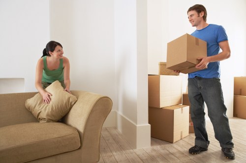 The image depicts a young man with light skin, wearing a blue t-shirt and dark jeans, standing inside a residential property in Greenwich, holding a medium-sized cardboard box. He is smiling and appears to be in the process of moving or packing. To his right, there are several other packed cardboard boxes of various sizes, some stacked on top of each other near the wall, indicating a household packing activity. In the foreground, a young woman with light skin, wearing a green sleeveless top, is seated on a beige sofa, holding a large beige cushion, and looking at the man with a friendly expression. The room features light-colored wood flooring and plain cream walls, with natural lighting illuminating the space. The scene captures an interior moment of house moving preparations, reflecting the house removals service context in Greenwich, with no other furniture or objects visible, focusing on the interaction and packing process typical of residential relocations in the area.
