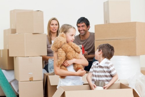 A family of four preparing for a house move indoors, surrounded by numerous cardboard moving boxes of various sizes, some stacked on top of each other and others open with items visible inside. The young girl, wearing a fluffy beige coat, is holding a teddy bear, while a boy in a striped shirt stands inside an open box holding a small metallic object. Behind them, a woman with long blonde hair and a man with dark hair and a smile are visible, attentively watching the children. The scene is set in a bright, well-lit room with a neutral wall background, and the boxes appear to be packed and ready for relocation, indicating a household in the process of moving, typical of local removals in the Marks Gate area.