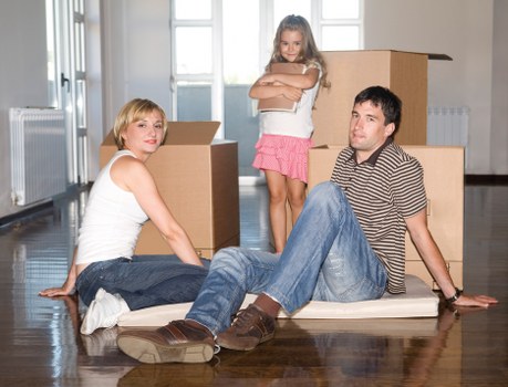 A family of three in a bright, modern room with large windows and wooden flooring, surrounded by open cardboard boxes for moving. The woman, with short blonde hair, is seated on the floor wearing a white tank top and blue jeans, looking directly at the camera. The man, with dark hair, is sitting on the floor with his legs extended, wearing a striped shirt and blue jeans, appearing relaxed. The young girl, with long blonde hair, is standing behind them, wearing a pink skirt and a white top, with crossed arms and a confident expression. The scene suggests an active packing or unpacking process as part of a house move, which is typical for house removals services in London or surrounding areas. The background shows a residential space with radiators, a window, and natural daylight illuminating the scene, emphasizing the domestic context of the relocation activity. Watson Rremovals London is known for assisting families with furniture and household item relocations within the local postcode area.