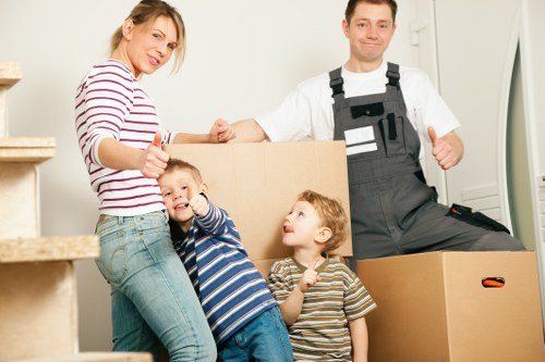 A family with a woman, two young children, and a man in a white t-shirt and black overalls, standing inside a residential property in White City, preparing to move. The woman, wearing a striped long-sleeve top and jeans, is giving a thumbs-up while holding a large cardboard box. One child, dressed in a blue and white striped shirt, is smiling and leaning against the woman, giving a thumbs-up with his hand. The other child, wearing a grey and white striped shirt, looks up at the woman. Behind them, there are several cardboard moving boxes stacked and on the floor, some partially opened. The setting appears to be a bright, clean interior with neutral-toned walls and a staircase to the left, indicating a typical home environment. The scene captures a moment of preparation and positivity associated with house removals, reflecting the services of Watson Removals London in the White City area, with household furniture or belongings such as boxes and childrenâ€™s toys visible, all arranged to convey a straightforward and organized moving process.