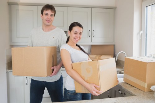 A man and a woman stand in a bright, modern kitchen with light grey cabinetry and a stainless steel sink, both holding cardboard boxes filled with household belongings. The man, dressed in a light grey sweater and dark jeans, stands behind the woman, who is wearing a white t-shirt and dark jeans, smiling gently at the camera. The womanâ€™s box appears to contain small items, while the manâ€™s box is empty or not fully visible. Several additional cardboard boxes are stacked on the kitchen counter to the right, some sealed with packing tape. The scene is well-lit by natural light coming through a large window on the right side, illuminating the space and emphasizing the clean, organized environment typical of house removals in Stockwell. This scene captures the process of packing and preparing household items for a relocation, consistent with professional removals service activities offered by Watson Removals London in the London Borough of Lambeth.