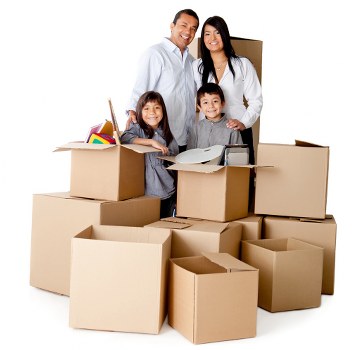 A family of four, including a man and woman, along with two children, is standing inside a room filled with unpacked cardboard boxes of various sizes, some open and filled with household items such as books, toys, and small appliances. The family members are smiling and appear to be in the process of moving, with several boxes stacked around them. The background features a plain white wall, and the scene is well-lit, highlighting the textures of the cardboard boxes and the household belongings. The setting suggests a residential interior typical of a home preparing for or completing a house move in Clapham, with Watson Removals London providing professional removal services. The arrangement of boxes and the presence of personal items reflect the typical activities associated with house removals and relocation, emphasizing a professional and organized move.