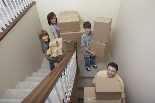 Four individuals, including two children and two adults, are positioned in a narrow staircase of a residential property, preparing for a house move. The staircase has a wooden handrail on the left and white spindles, with beige carpeted steps. The children, a girl with long red hair and a boy with short dark hair, are holding medium-sized cardboard boxes; the girl has a teddy bear in her box, and the boy is carrying a box with another box stacked beside him on the stairs. One of the adults, a woman with shoulder-length dark hair, is standing on the landing near a large cardboard box, while the other adult, a man with short dark hair, is at the bottom of the stairs holding a box. The background shows plain cream-colored walls and a wooden window ledge with vertical blinds. The scene appears to be set during daytime with natural light illuminating the space, indicative of a typical house relocation undertaken by Watson Removals London in the Belvedere area, showcasing a family engaged in packing or unpacking belongings during a home move.